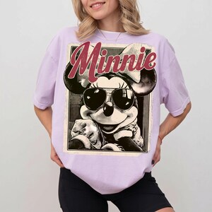 May include: A light purple t-shirt featuring a black and white graphic of Minnie Mouse wearing sunglasses. The word "Minnie" is printed in maroon script above the graphic. The shirt has short sleeves and a relaxed fit.