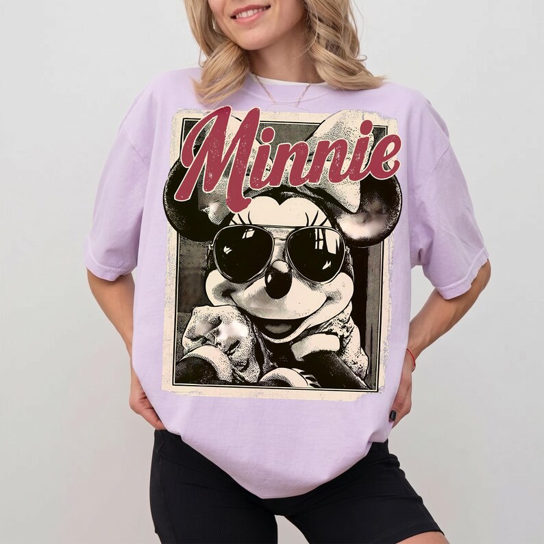May include: A light purple t-shirt featuring a black and white graphic of Minnie Mouse wearing sunglasses. The word "Minnie" is printed in maroon script above the graphic. The shirt has short sleeves and a relaxed fit.