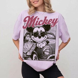 May include: A light purple t-shirt featuring a graphic of Mickey Mouse wearing sunglasses and leaning against a car. The word "Mickey" is printed above the graphic in a maroon, retro font. The shirt is a relaxed fit.