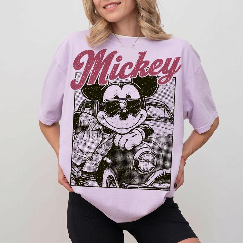 May include: A light purple t-shirt featuring a graphic of Mickey Mouse wearing sunglasses and leaning against a car. The word "Mickey" is printed above the graphic in a maroon, retro font. The shirt is a relaxed fit.
