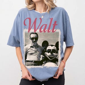May include: A pale blue t-shirt with a retro image of Walt Disney and Mickey Mouse, both sporting sunglasses. The word "Walt" is printed in a faded red font above the image. The shirt has short sleeves and a crew neck.