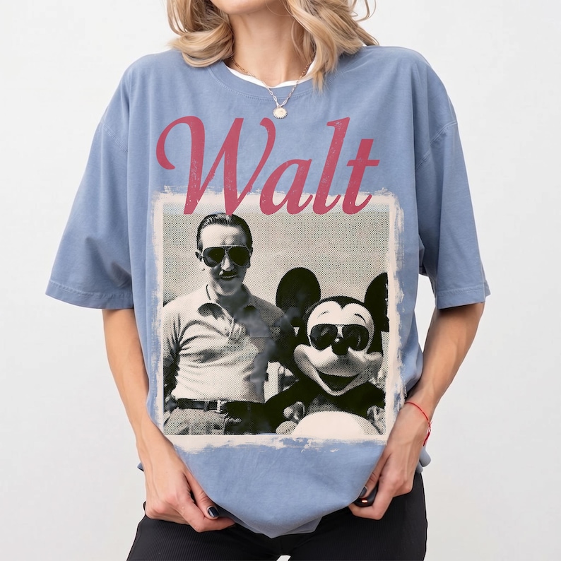May include: A pale blue t-shirt with a retro image of Walt Disney and Mickey Mouse, both sporting sunglasses. The word "Walt" is printed in a faded red font above the image. The shirt has short sleeves and a crew neck.