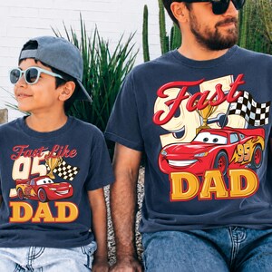 May include: Two navy blue t-shirts with a graphic of a red race car, a trophy, and a checkered flag. The text on the shirts reads "Fast Like 95 Dad" in yellow and red. One shirt is worn by a child, the other by an adult.