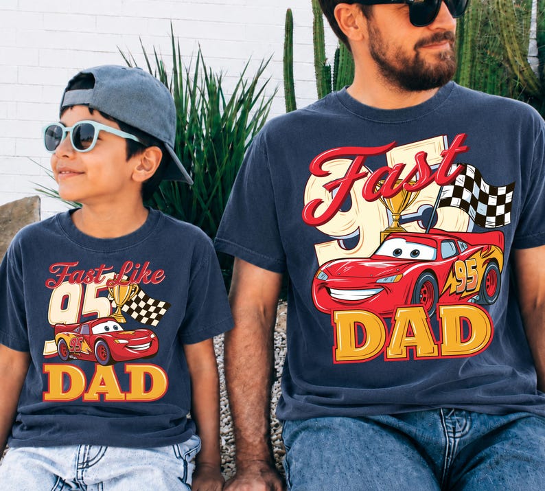 May include: Two navy blue t-shirts with a graphic of a red race car, a trophy, and a checkered flag. The text on the shirts reads "Fast Like 95 Dad" in yellow and red. One shirt is worn by a child, the other by an adult.