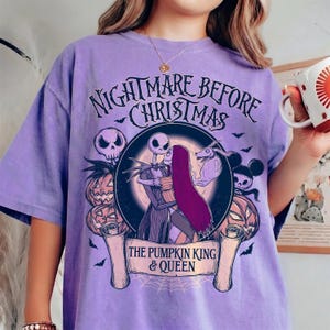 May include: A lilac t-shirt with a graphic from "The Nightmare Before Christmas." The design features Jack and Sally embracing, with pumpkins, skulls, and bats. The text reads "Nightmare Before Christmas" and "The Pumpkin King & Queen."