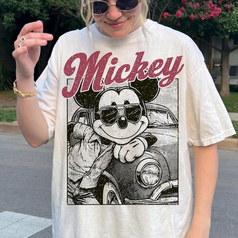 May include: Cream-colored t-shirt featuring a graphic of Mickey Mouse wearing sunglasses and leaning on a vintage car. The word "Mickey" is printed in a burgundy, retro font above the graphic.