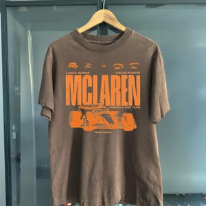 May include: Brown t-shirt featuring the McLaren Formula One Team logo in orange. The design includes the names Lando Norris and Oscar Piastri, along with a graphic of a race car. The words "Team Papaya" are also visible.