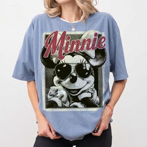 May include: A light blue t-shirt featuring a graphic of Minnie Mouse wearing sunglasses. The word "Minnie" is printed in a burgundy script across the top of the graphic. The t-shirt has short sleeves and a relaxed fit.