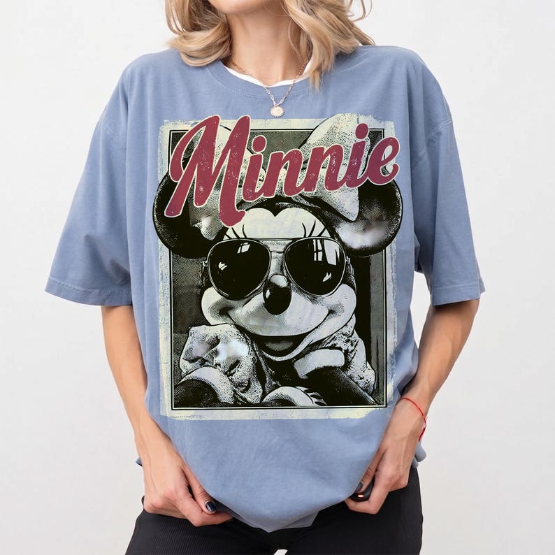 May include: A light blue t-shirt featuring a graphic of Minnie Mouse wearing sunglasses. The word "Minnie" is printed in a burgundy script across the top of the graphic. The t-shirt has short sleeves and a relaxed fit.