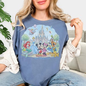 May include: Blue t-shirt featuring a colorful illustration of Disney characters including Mickey Mouse, Minnie Mouse, Donald Duck, and Ariel. The graphic includes a castle and the text "Magic Kingdom" and "Walt Disney World".