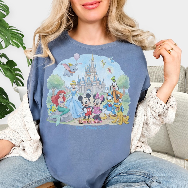 May include: Blue t-shirt featuring a colorful illustration of Disney characters including Mickey Mouse, Minnie Mouse, Donald Duck, and Ariel. The graphic includes a castle and the text "Magic Kingdom" and "Walt Disney World".