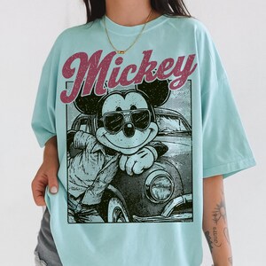 May include: Light blue t-shirt featuring a graphic of Mickey Mouse wearing sunglasses and leaning against a vintage car. The word "Mickey" is printed above the graphic in a retro font. The shirt has a relaxed fit.