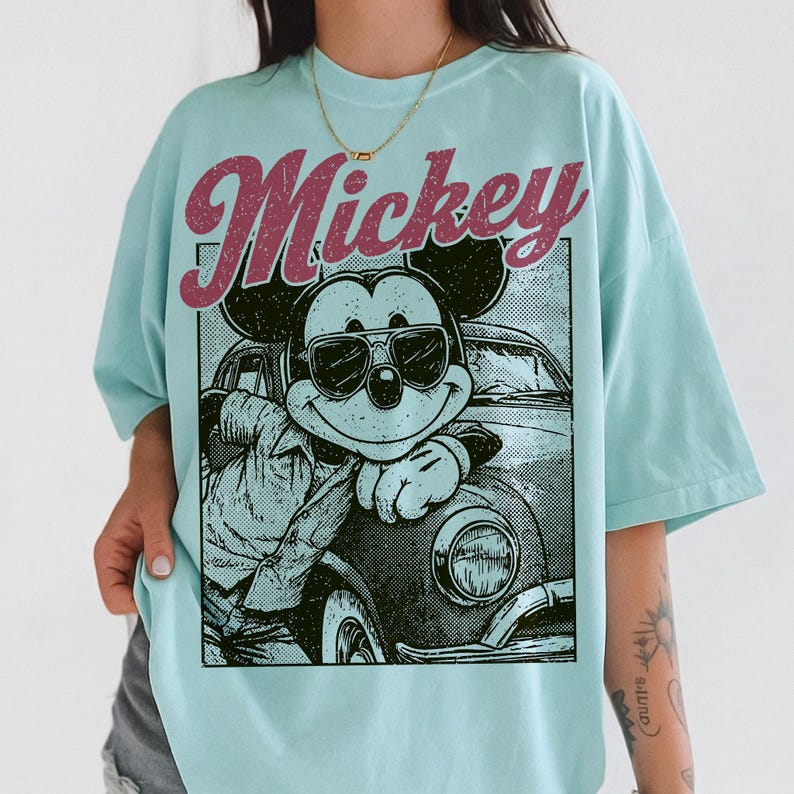 May include: Light blue t-shirt featuring a graphic of Mickey Mouse wearing sunglasses and leaning against a vintage car. The word "Mickey" is printed above the graphic in a retro font. The shirt has a relaxed fit.