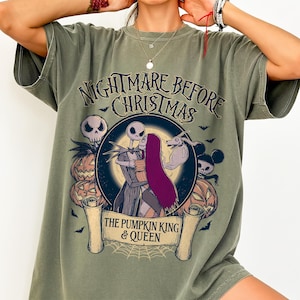 May include: Olive green t-shirt with a graphic from "The Nightmare Before Christmas." The design features Jack and Sally embracing, surrounded by pumpkins, bats, and the text "The Pumpkin King & Queen."