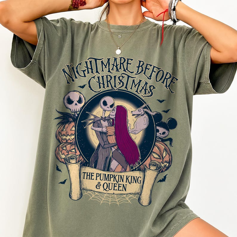 May include: Olive green t-shirt with a graphic from "The Nightmare Before Christmas." The design features Jack and Sally embracing, surrounded by pumpkins, bats, and the text "The Pumpkin King & Queen."