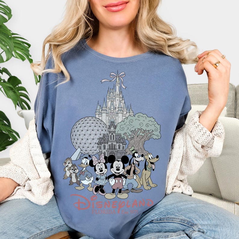 K&ouml;nnte beinhalten: Hellblaues T-Shirt mit einer Grafik von Disney-Figuren, darunter Micky Maus, Minnie Maus und Goofy, vor dem Cinderella-Schloss. Der Text "Disneyland Florida" ist unter den Figuren aufgedruckt.