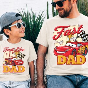May include: Two matching beige t-shirts with the text "Fast Like 95 Dad" and a cartoon image of a red race car. The shirts are worn by a father and child, both wearing sunglasses.