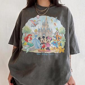May include: Dark gray t-shirt featuring a colorful graphic of Disney characters including Mickey and Minnie Mouse, Cinderella, Ariel, and Dumbo in front of a castle. The text "Magic Kingdom" and "Walt Disney World" are also visible.