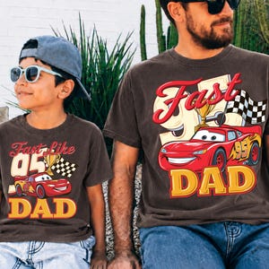 May include: Two brown t-shirts with matching graphics. One shirt says "Fast like 05 DAD" with a red race car and trophy. The other shirt says "Fast DAD" with a red race car, trophy, and checkered flag. Both shirts have yellow and red text.