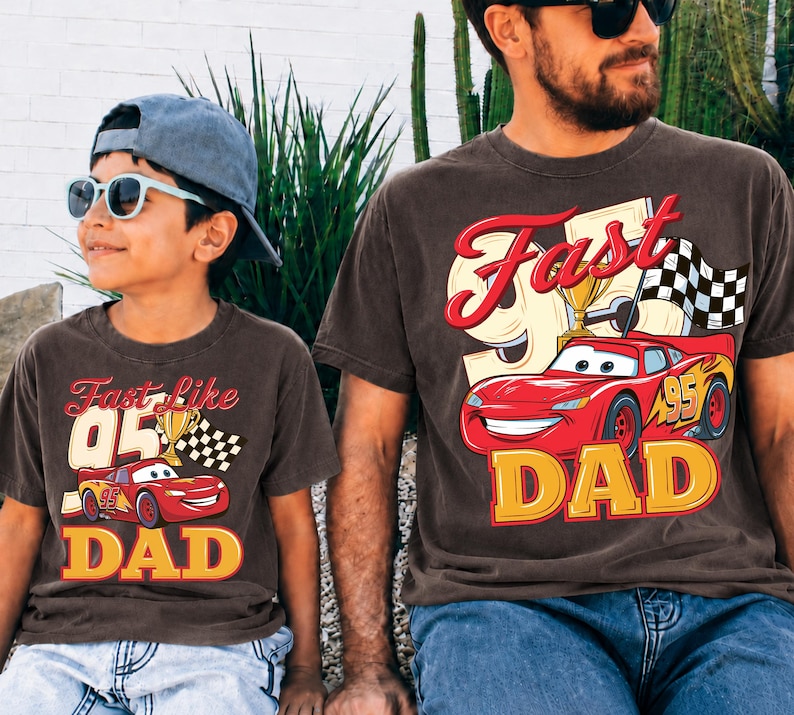 May include: Two brown t-shirts with matching graphics. One shirt says "Fast like 05 DAD" with a red race car and trophy. The other shirt says "Fast DAD" with a red race car, trophy, and checkered flag. Both shirts have yellow and red text.