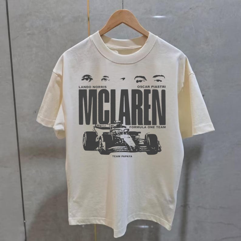 May include: Cream-colored t-shirt featuring a black graphic of a Formula One race car and the word "MCLAREN" in bold letters. The text "LANDO NORRIS" and "OSCAR PIASTRI" are above the word "MCLAREN". The words "FORMULA ONE TEAM" and "TEAM PAPAYA" are also printed on the shirt.