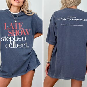 May include: A dark blue t-shirt featuring the text "The Late Show with Stephen Colbert" in red and white. The back of the shirt has the text "05.21.2026 The Night The Laughter Died" in white. The shirt is a short-sleeved, oversized fit.
