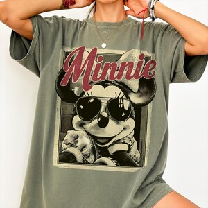 May include: Olive green t-shirt featuring a graphic of Minnie Mouse wearing sunglasses. The word "Minnie" is printed in a burgundy script across the top of the graphic. The shirt has short sleeves and a relaxed fit.