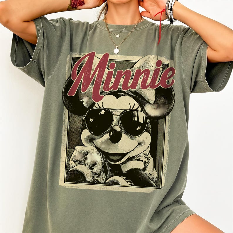 May include: Olive green t-shirt featuring a graphic of Minnie Mouse wearing sunglasses. The word "Minnie" is printed in a burgundy script across the top of the graphic. The shirt has short sleeves and a relaxed fit.