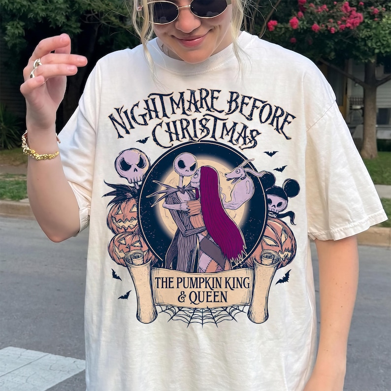 May include: Cream-coloured t-shirt featuring a graphic from "The Nightmare Before Christmas." The design includes Jack Skellington and Sally embracing, surrounded by pumpkins, bats, and the text "The Pumpkin King & Queen."