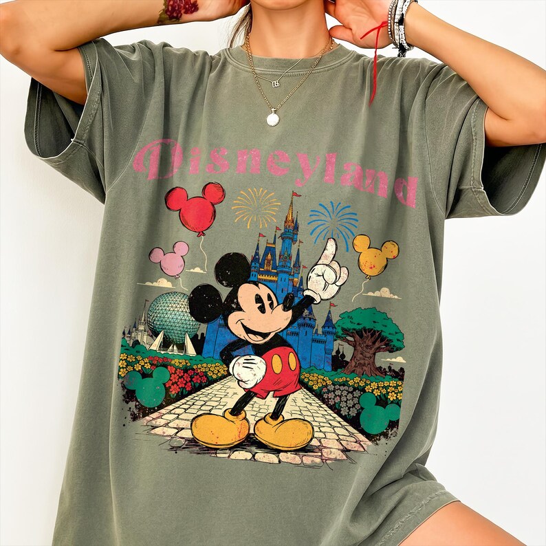 Puede incluir: Camiseta verde oliva con un gr&aacute;fico de Mickey Mouse se&ntilde;alando un castillo, con la palabra "Disneyland" en rosa. El dise&ntilde;o incluye globos, fuegos artificiales y un paisaje colorido. La camiseta tiene mangas cortas y un corte holgado.