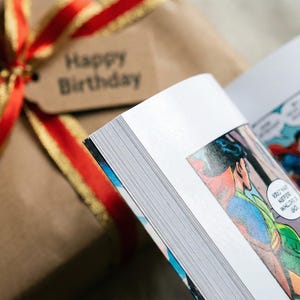 May include: A wrapped gift with a red and gold ribbon and a tag that says "Happy Birthday" next to an open comic book. The comic book features colorful illustrations of superheroes.