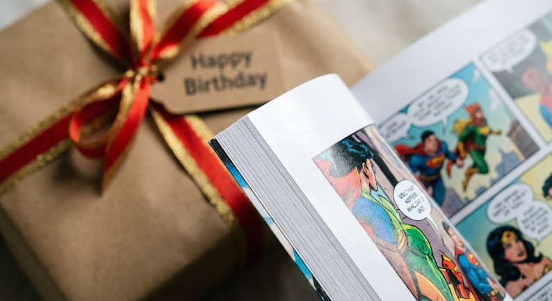 May include: A wrapped gift with a red and gold ribbon and a tag that says "Happy Birthday" next to an open comic book. The comic book features colorful illustrations of superheroes.