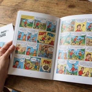 May include: An open comic book with colorful panels featuring a cartoon character in various scenes. The cover of the book, titled "LES 50 ANS DE MARC: L'AVENTURE", is visible next to it. The book is on a wooden surface.