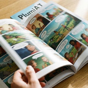 Puede incluir: Cómic abierto con paneles coloridos y bocadillos. La portada del libro, titulado "PlumiAT", es visible en el fondo. Las páginas se están pasando, revelando arte secuencial con personajes en varias escenas.
