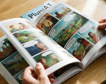 May include: Open comic book with colorful panels and speech bubbles. The cover of the book, titled "PlumiAT", is visible in the background. The pages are being turned, revealing sequential art with characters in various scenes.