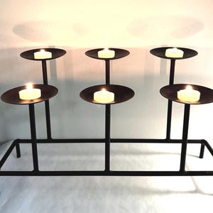 May include: A black metal candelabra featuring six circular platforms arranged at two different heights. Each platform holds a single lit white tea light candle. The structure is supported by a rectangular base frame with four thin legs.