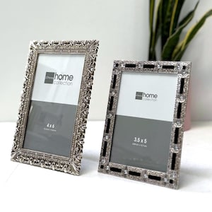 May include: Two silver-colored picture frames with ornate detailing. One frame is 4x6 inches, the other is 3.5x5 inches. Both frames have a white and gray interior and the text "home collection".