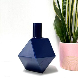 May include: A dark blue ceramic vase with a geometric, multi-faceted diamond shape and a narrow cylindrical neck. The surface has a smooth, glossy finish that reflects light, creating sharp edges and distinct angles across the body of the object.