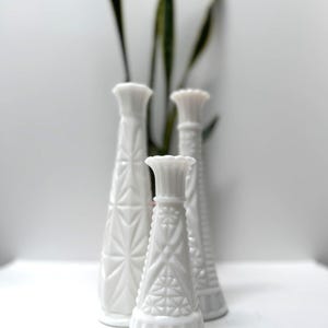 May include: Four white, textured glass vases of varying heights. The vases have a geometric pattern and a flared top. Green leaves are visible in the background. The vases are arranged on a white surface.