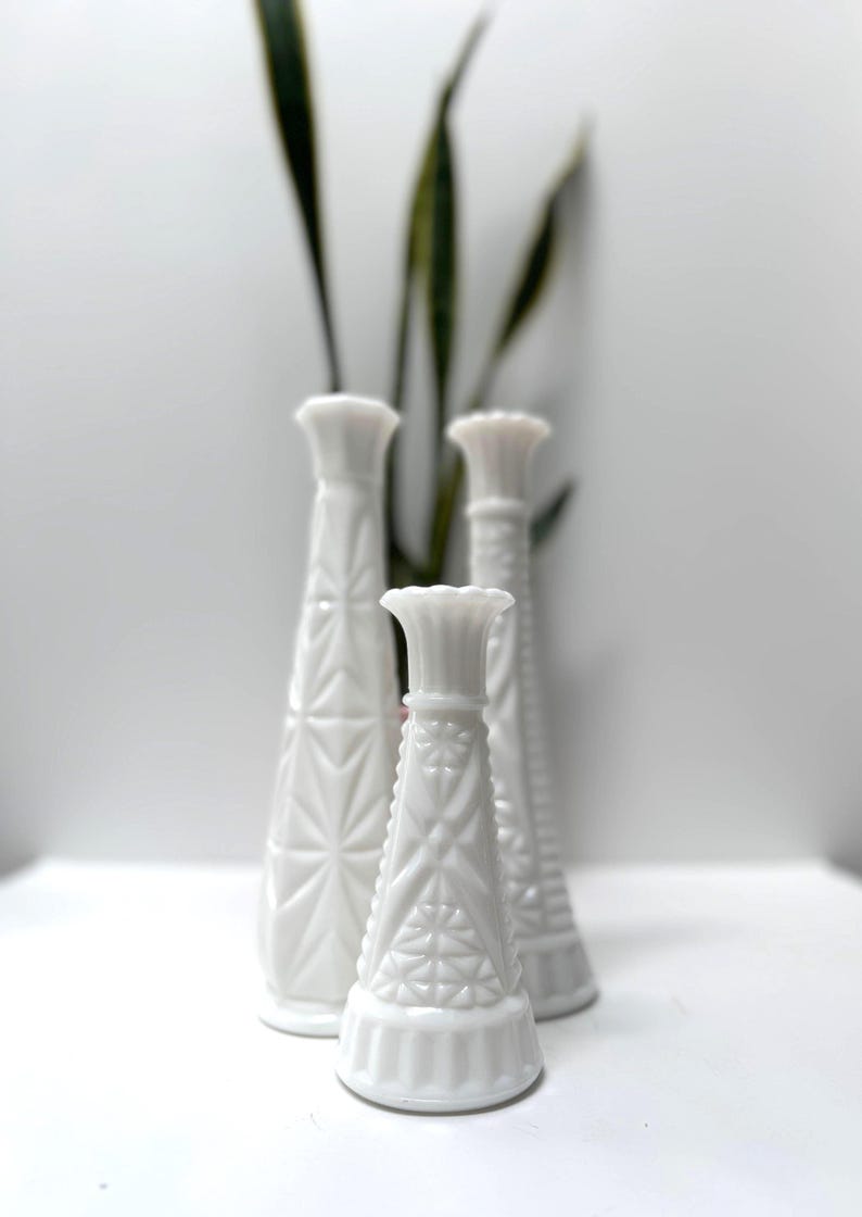 May include: Four white, textured glass vases of varying heights. The vases have a geometric pattern and a flared top. Green leaves are visible in the background. The vases are arranged on a white surface.