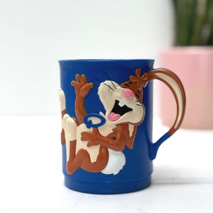 Peut inclure: Mug en céramique bleue avec un personnage de dessin animé marron, blanc et rose. Le personnage est dans une pose ludique, avec une grande bouche ouverte et un anneau bleu. La tasse a une anse marron.