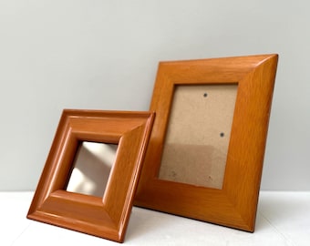 May include: Two wooden picture frames in a warm, reddish-brown hue. One frame is square, the other is rectangular. Both have a simple, classic design with a slightly glossy finish. The frames are empty, ready to display photos or artwork.