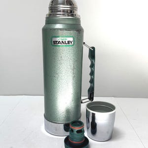 May include: A vintage green Stanley Aladdin thermos with a metallic finish, a dark green plastic side handle, and a silver-colored cap. The thermos features a white label with the text Aladdin STANLEY. A separate dark green stopper and a silver cup.