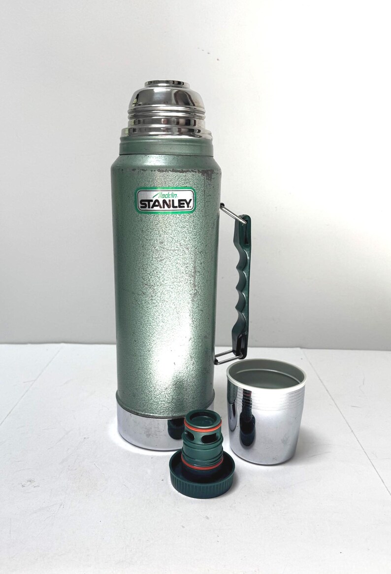 May include: A vintage green Stanley Aladdin thermos with a metallic finish, a dark green plastic side handle, and a silver-colored cap. The thermos features a white label with the text Aladdin STANLEY. A separate dark green stopper and a silver cup.
