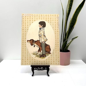 May include: Vintage illustration of a boy and a dog. The artwork features a boy in a coat and pants standing next to a brown dog. The background is a cream color with a repeating gold pattern. The artwork is displayed on a small, dark wooden stand.