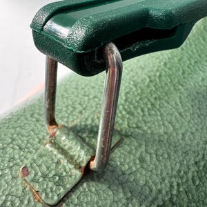 May include: A dark green plastic handle attached to a textured light green surface by a silver-colored metal loop. The metal loop connects to a small, rectangular, light green metal plate that shows signs of rust around the edges where it meets the surface.