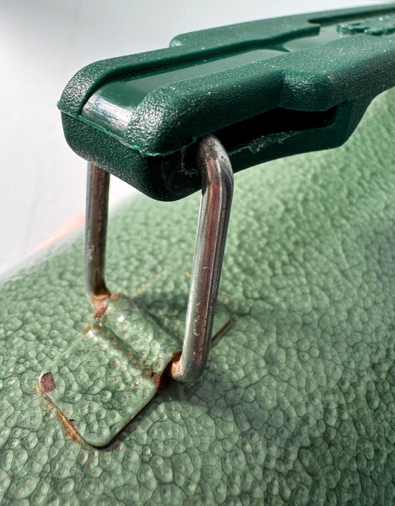 May include: A dark green plastic handle attached to a textured light green surface by a silver-colored metal loop. The metal loop connects to a small, rectangular, light green metal plate that shows signs of rust around the edges where it meets the surface.