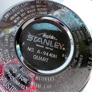 May include: A circular, metallic base of a Stanley vacuum bottle with engraved text including 'Aladdin', 'Stanley', 'No. A-944DH', and 'Quart'. The surface features instructions for preheating with hot water and cooling with cold water.