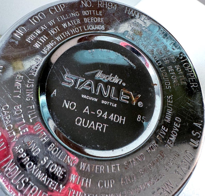 May include: A circular, metallic base of a Stanley vacuum bottle with engraved text including 'Aladdin', 'Stanley', 'No. A-944DH', and 'Quart'. The surface features instructions for preheating with hot water and cooling with cold water.