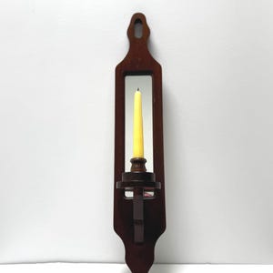May include: A dark brown wooden wall sconce with a yellow candle. The sconce has a rectangular shape with a decorative cutout and a candle holder. The candle is a standard size and is centered in the sconce.